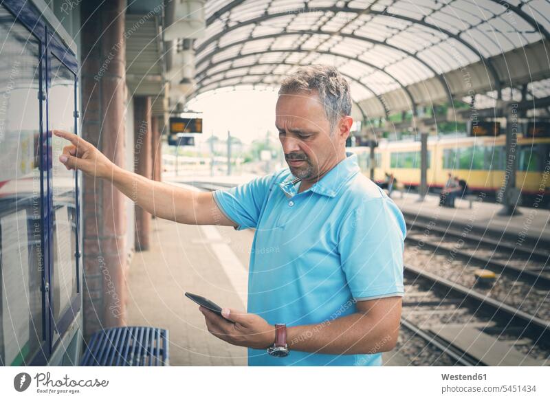 Mann mit Smartphone steht auf dem Bahnsteig vor dem Fahrplan Bahnhof Bahnhöfe Bahnhoefe Männer männlich Station Haltestelle Haltestellen Stationen Erwachsener