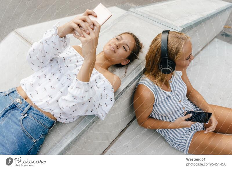 Zwei junge Frauen mit Mobiltelefonen und Kopfhörern in einem Skatepark Freundinnen Kopfhoerer Handy Handies Handys weiblich Skateboardpark Skateboard-Park