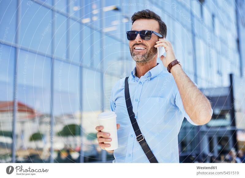 Fröhlicher Geschäftsmann spricht am Telefon und hält Kaffee im Freien lächeln telefonieren anrufen Anruf telephonieren Handy Mobiltelefon Handies Handys