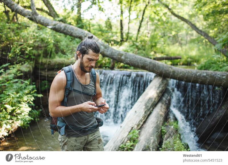 Junger Mann hält Handy an einem Wasserfall im Wald Mobiltelefon Handies Handys Mobiltelefone Wanderer wandern Wanderung Männer männlich Telefon telefonieren