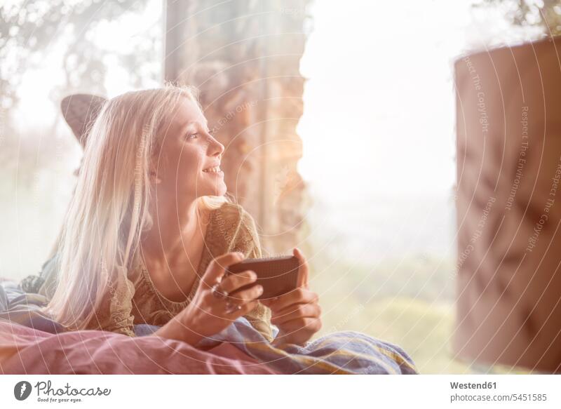Porträt einer lächelnden jungen Frau, die mit ihrem Handy auf dem Bett liegt und durch das Fenster schaut weiblich Frauen Erwachsener erwachsen Mensch Menschen
