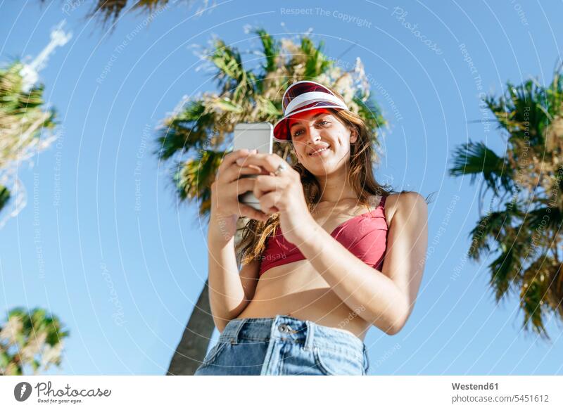 Junge Frau mit Sonnenblende und Bikini telefoniert stehen stehend steht lächeln Handy Mobiltelefon Handies Handys Mobiltelefone weiblich Frauen Telefon