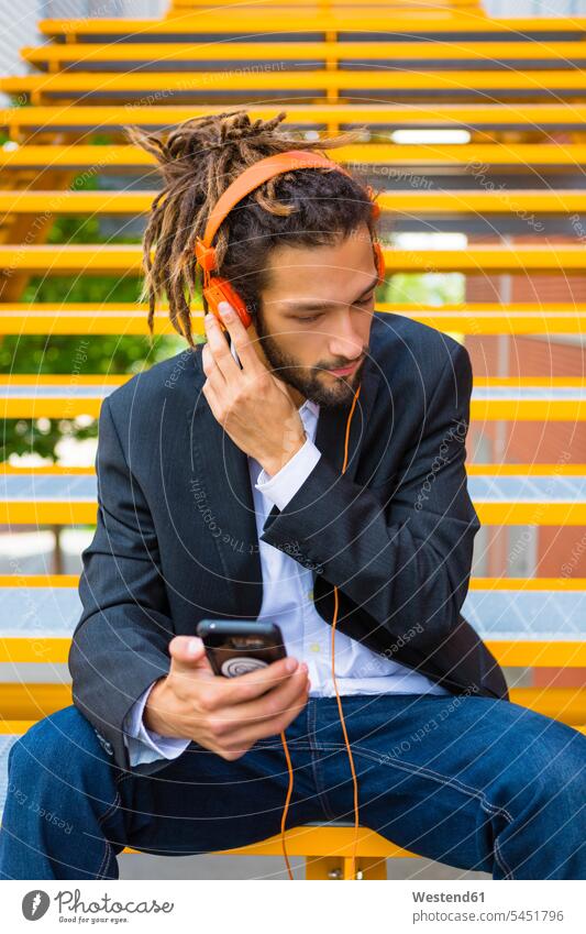 Junger Geschäftsmann mit Dreadlocks sitzt auf der Treppe und hört mit Kopfhörer und Handy Musik Kopfhoerer Businessmann Businessmänner Geschäftsmänner