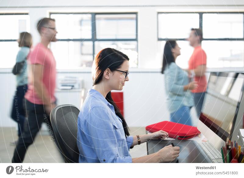 Mitarbeiterin, die an ihrem Computer arbeitet, während andere Mitarbeiter vorbeilaufen Büro Office Büros Arbeitsplatz Arbeitsstätte Arbeitstelle Kollegen