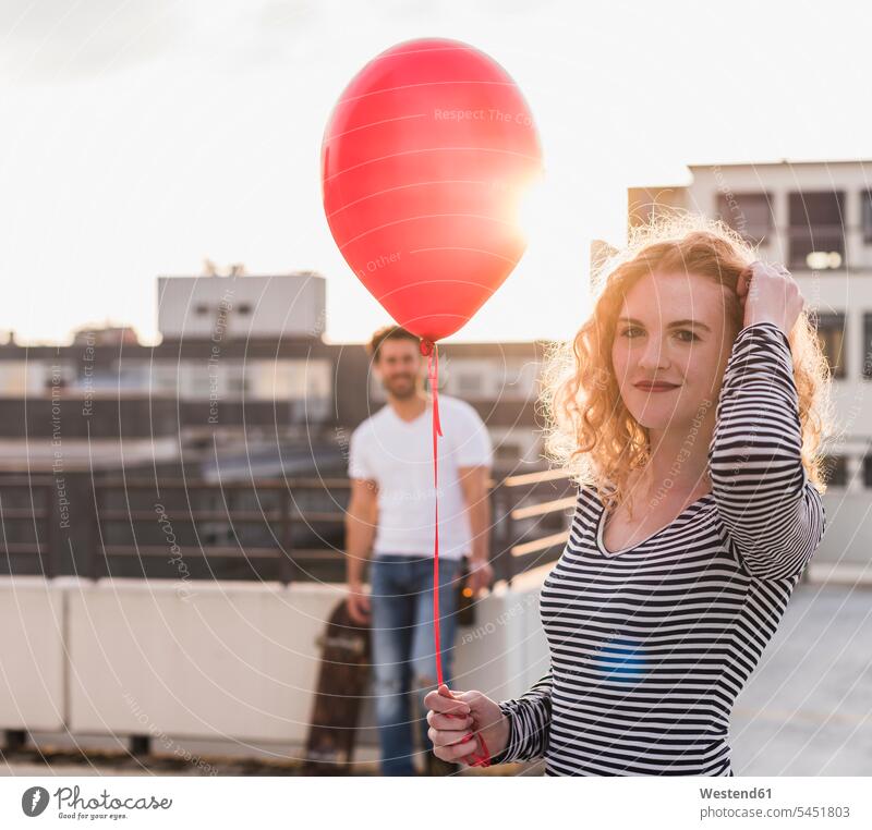 Porträt eines lächelnden jungen Wo-Mannes mit rotem Ballon auf der Dachterrasse bei Sonnenuntergang Luftballons Ballons Dachterrassen Luftballone roter rotes
