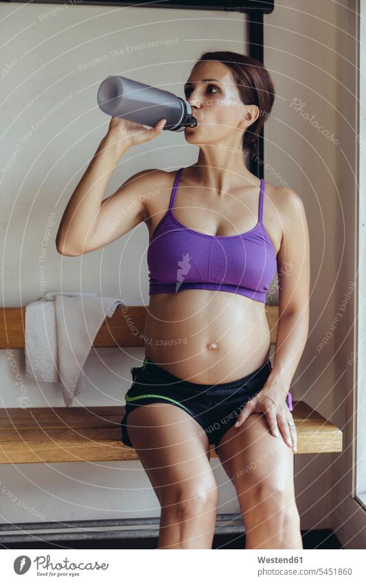 Sportliche Schwangere sitzt im Umkleideraum und trinkt aus der Flasche Frau weiblich Frauen sitzen sitzend trinken Wasserflasche Flaschen Wasserflaschen