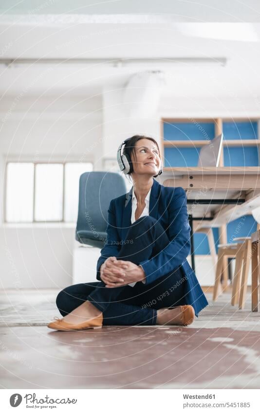 Lächelnde Geschäftsfrau, die auf dem Boden in einem Loft sitzt und mit Kopfhörern Musik hört Kopfhoerer Frau weiblich Frauen Geschäftsfrauen Businesswomen