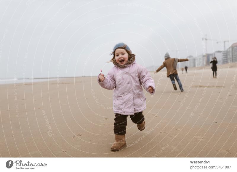 Glückliches kleines Mädchen mit Lolly, das im Winter am Strand läuft, während ihr Bruder im Hintergrund spielt glücklich glücklich sein glücklichsein spielen
