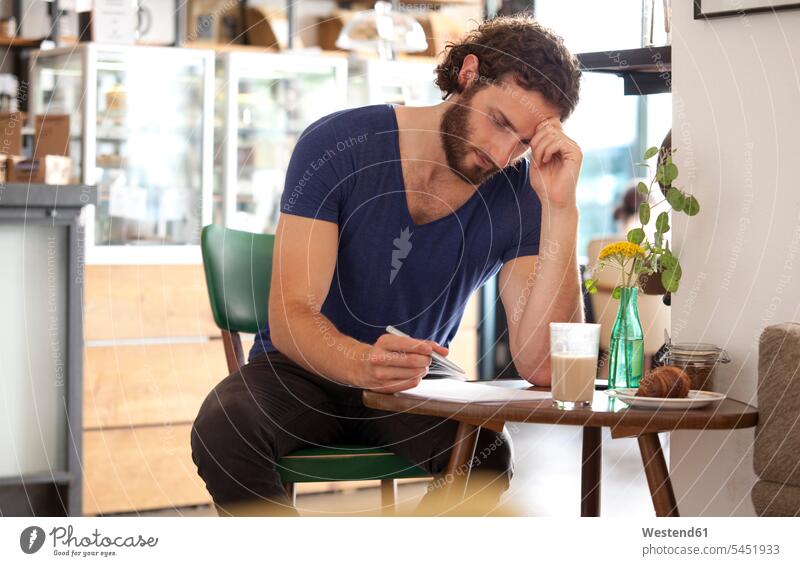 Nachdenklicher junger Mann sitzt in einem Café und schreibt einen Brief schreiben aufschreiben notieren schreibend Schrift Cafe Kaffeehaus Bistro Cafes