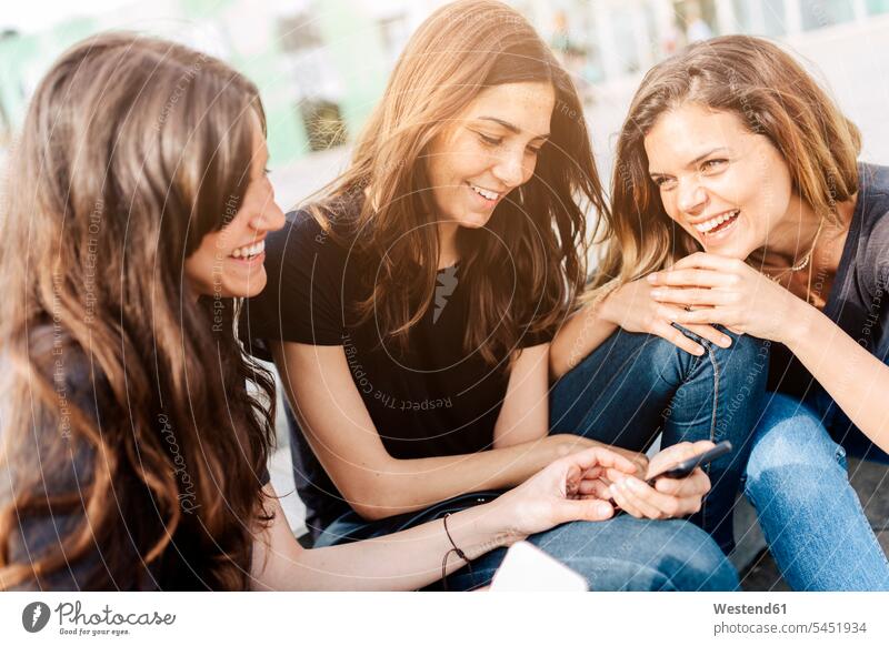 Drei glückliche junge Frauen sitzen im Freien und schauen auf ihr Handy lächeln Freundinnen Mobiltelefon Handies Handys Mobiltelefone Freunde Freundschaft