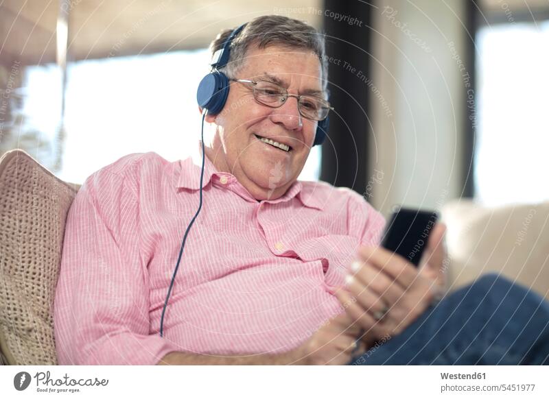 Lächelnder älterer Mann mit Kopfhörern, der zu Hause Musik hört Kopfhoerer Sofa Couches Liege Sofas hören hoeren Zuhause daheim Männer männlich lächeln Senior