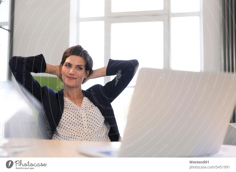 Glückliche Geschäftsfrau sitzt im Büro und schaut mit den Händen hinter dem Kopf auf den Laptop lächeln glücklich glücklich sein glücklichsein Notebook Laptops
