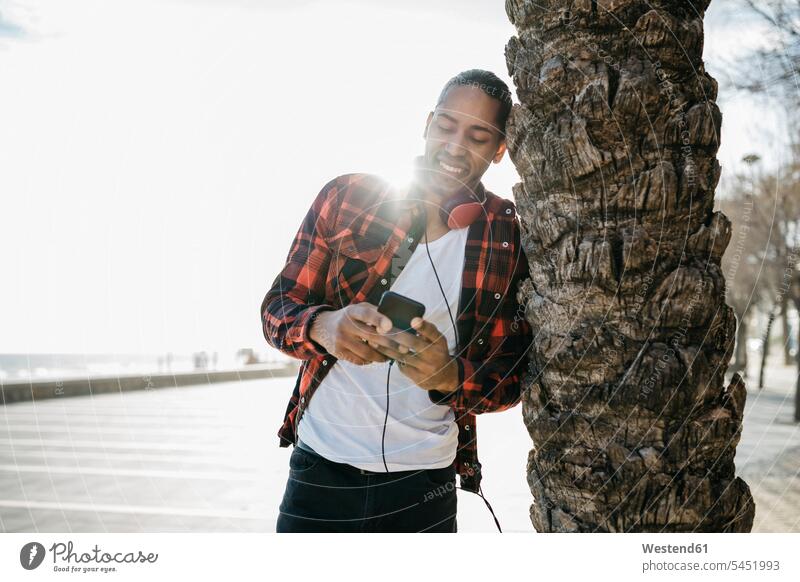 Spanien, lächelnder junger Mann mit Kopfhörern, der sich an einen Palmenstamm lehnt und auf sein Handy schaut Smartphone iPhone Smartphones Kopfhoerer Männer