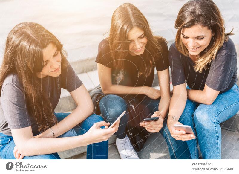 Drei lächelnde junge Frauen sitzen im Freien und schauen auf Mobiltelefone Freundinnen Handy Handies Handys Freunde Freundschaft Kameradschaft Telefon