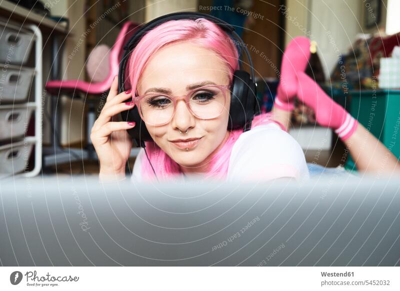 Junge Frau mit rosa Haaren hört zu Hause Musik über ihren Laptop weiblich Frauen lächeln Kopfhörer Kopfhoerer Brille Brillen Erwachsener erwachsen Mensch