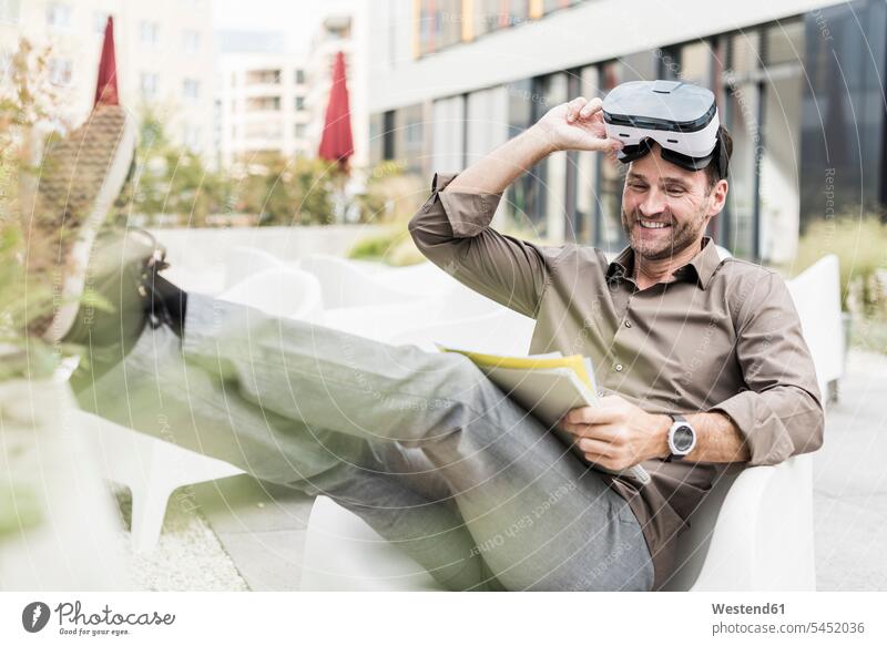 Lächelnder Mann mit Virtual-Reality-Brille und Dokumenten auf der Terrasse sitzend Geschäftsmann Businessmann Businessmänner Geschäftsmänner lächeln