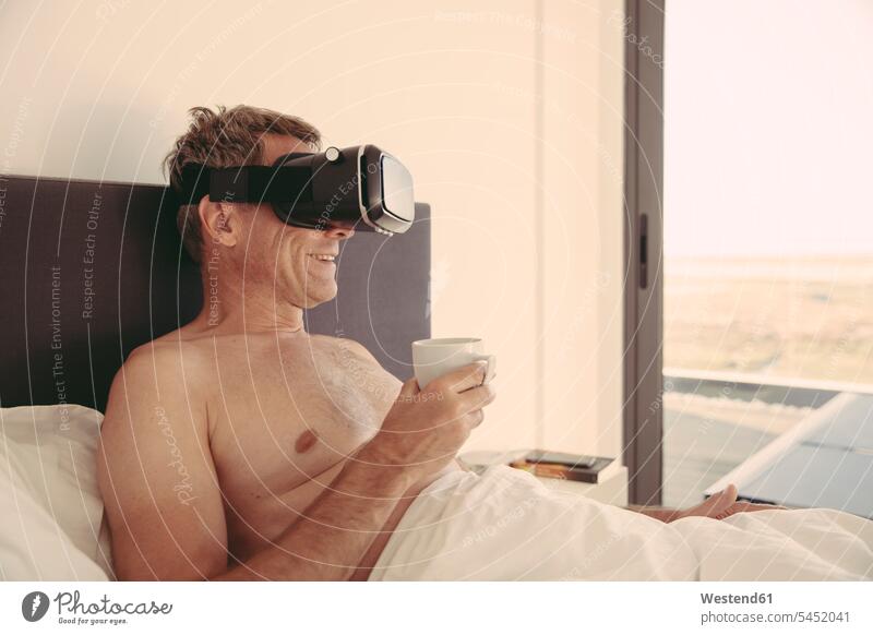 Mann mit VR-Brille im Bett mit einer Tasse Kaffee lächeln Brillen Männer männlich Wohnung Mietwohnungen wohnen Betten Virtuelle Realität Virtuelle Realitaet