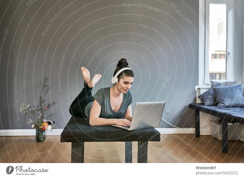 Junge Frau mit Kopfhörern, die mit einem Laptop im Wohnzimmer liegt Kopfhoerer liegen liegend Notebook Laptops Notebooks weiblich Frauen Computer Rechner
