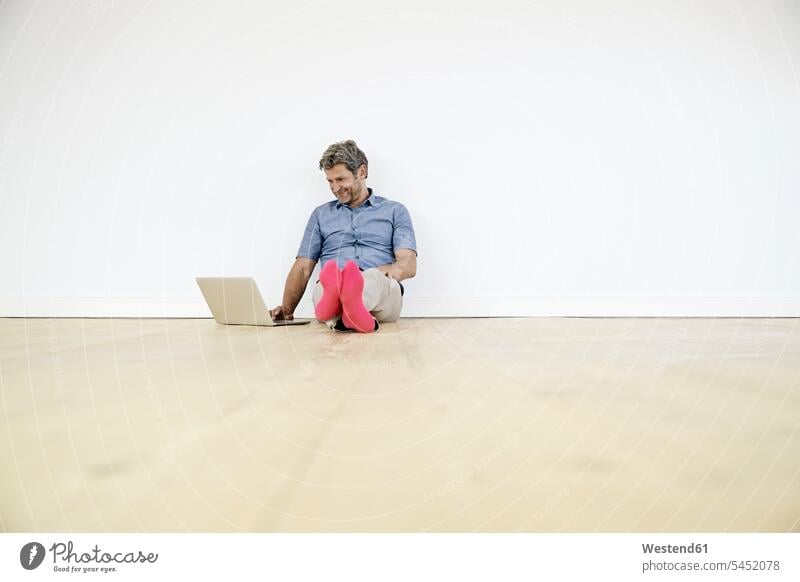 Älterer Mann sitzt auf dem Boden in einem leeren Raum und benutzt einen Laptop Männer männlich auf dem Boden sitzen auf dem Boden sitzend Zuhause zu Hause
