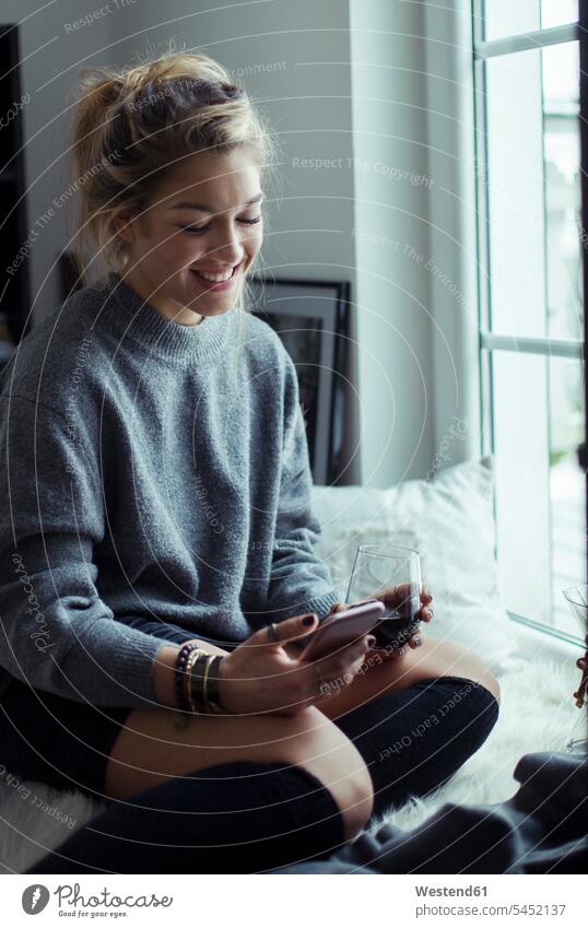 Lächelnde junge Frau mit einer Tasse Kaffee, die sich zu Hause auf dem Schaffell entspannt und auf ihr Handy schaut weiblich Frauen Smartphone iPhone