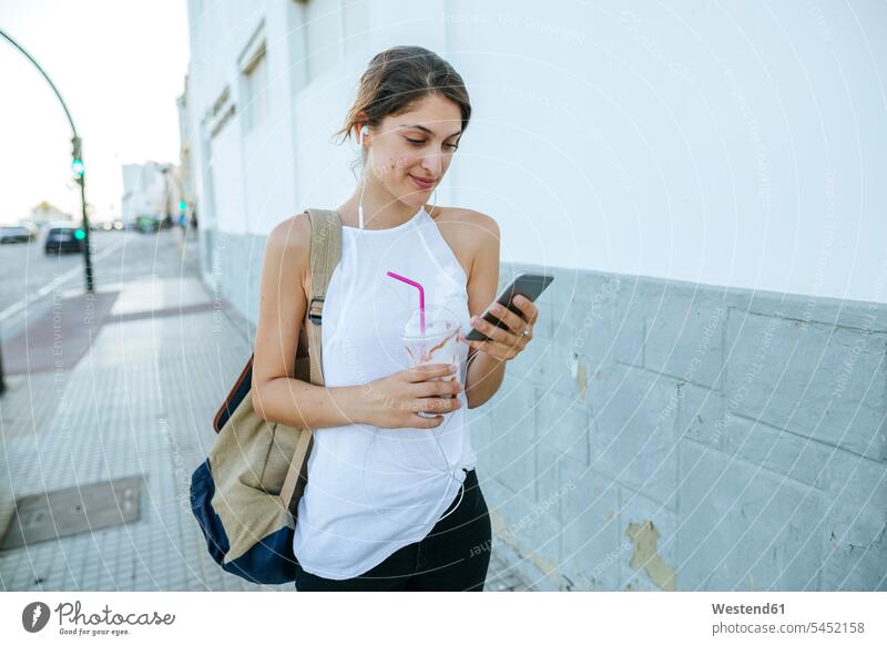 Junge Frau mit Handy und Smoothie geht die Straße entlang Smoothies Touristin weiblich Frauen Mobiltelefon Handies Handys Mobiltelefone Getränk Getraenk