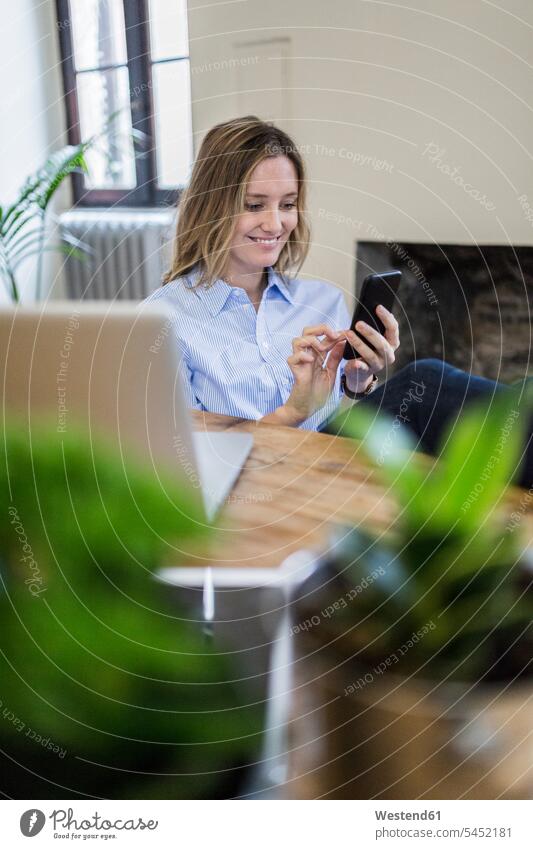 Lächelnde Frau sitzt zu Hause am Schreibtisch und kontrolliert ihr Handy Zuhause daheim weiblich Frauen sitzen sitzend lächeln Mobiltelefon Handies Handys