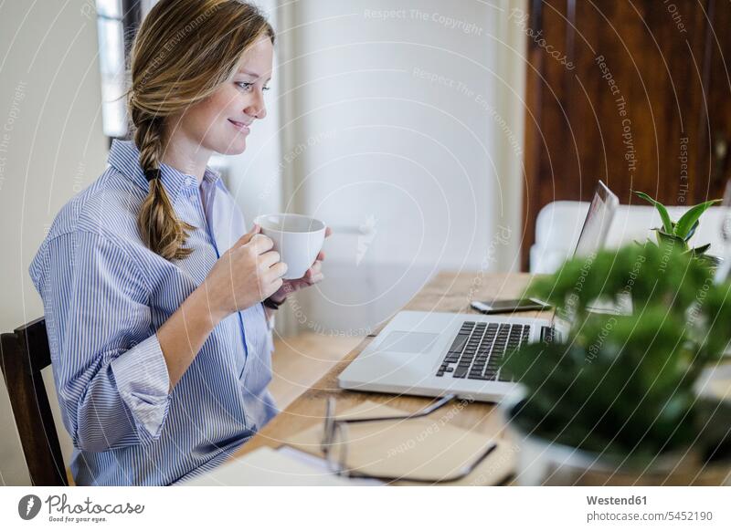 Lächelnde Frau sitzt am Schreibtisch mit Tasse Kaffee und Laptop Arbeitstisch Schreibtische sitzen sitzend lächeln Notebook Laptops Notebooks weiblich Frauen