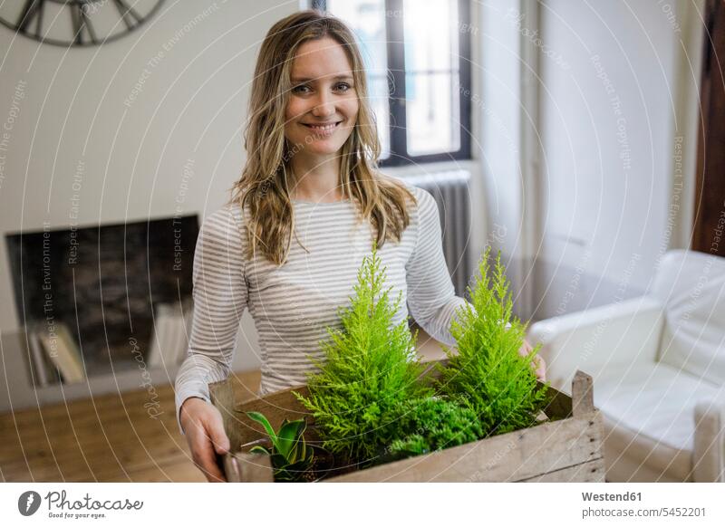 Porträt einer lächelnden Frau, die zu Hause eine Kiste mit Pflanzen trägt Portrait Porträts Portraits tragen transportieren Pflanzenwelt Flora weiblich Frauen