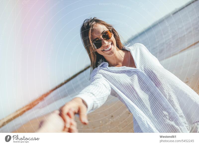 Porträt einer lächelnden jungen Frau mit Sonnenbrille, die die Hand am Strand hält weiblich Frauen Beach Straende Strände Beaches Portrait Porträts Portraits
