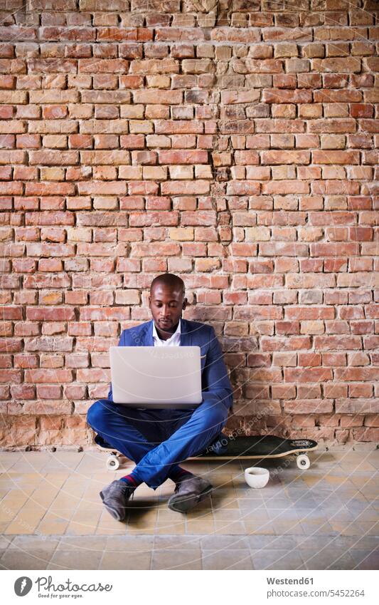 Geschäftsmann sitzt mit Laptop auf einem Longboard an einer Ziegelmauer Notebook Laptops Notebooks Businessmann Businessmänner Geschäftsmänner Skateboard