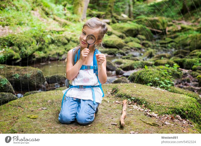 Kleines Mädchen mit Lupe, das auf einem Felsen im Wald kauert und eine Feder beobachtet weiblich Kind Kinder Kids Mensch Menschen Leute People Personen