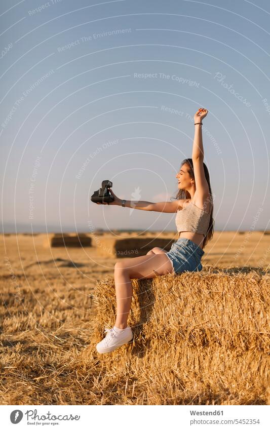 Junge Frau sitzt auf einem Strohballen und macht ein Selfie mit einer Sofortbildkamera Selfies weiblich Frauen Erwachsener erwachsen Mensch Menschen Leute