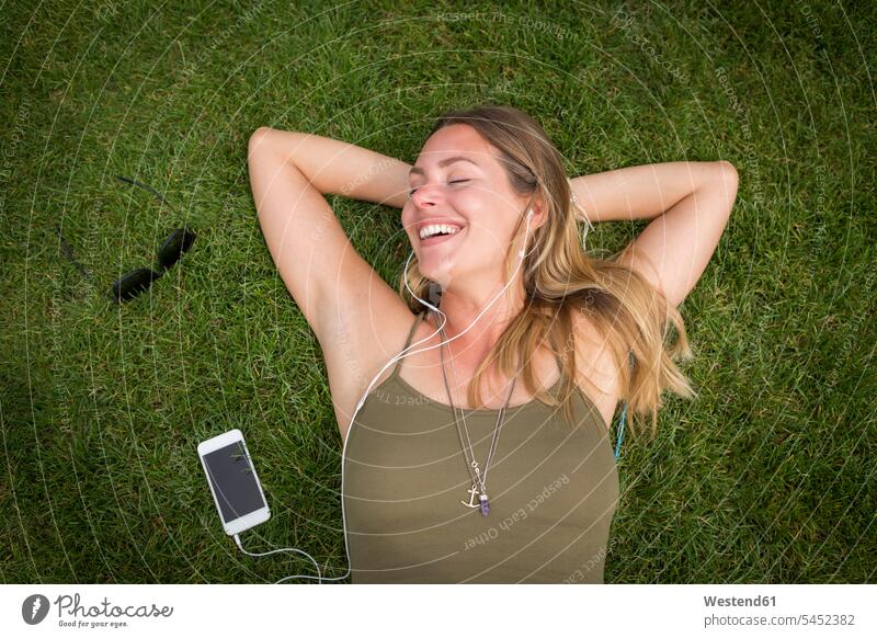 Glückliche junge Frau mit Kopfhörern entspannt auf dem Rasen Ohrhörer Portrait Porträts Portraits lächeln entspanntheit relaxt Park Parkanlagen Parks liegen