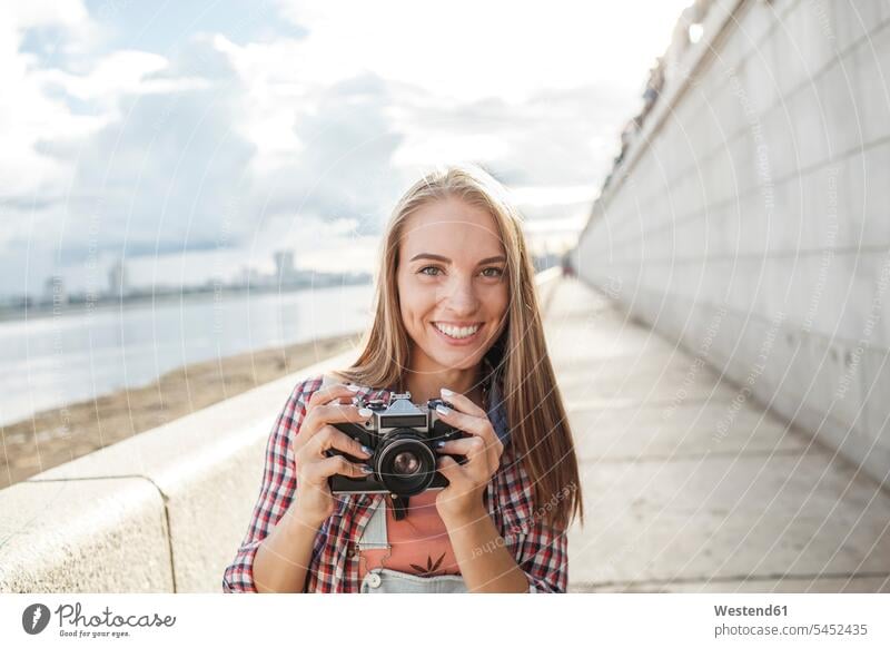 Lächelnde junge Frau mit einer Kamera am Flussufer Portrait Porträts Portraits Fotoapparat Fotokamera lächeln weiblich Frauen Fluesse Fluß Flüsse Erwachsener