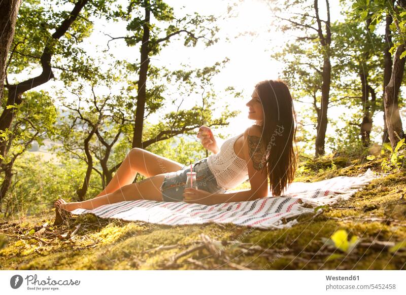 Lächelnde junge Frau im Wald auf einer Decke liegend mit Getränk und Apfel Forst Wälder lächeln entspannt entspanntheit relaxt weiblich Frauen Entspannung