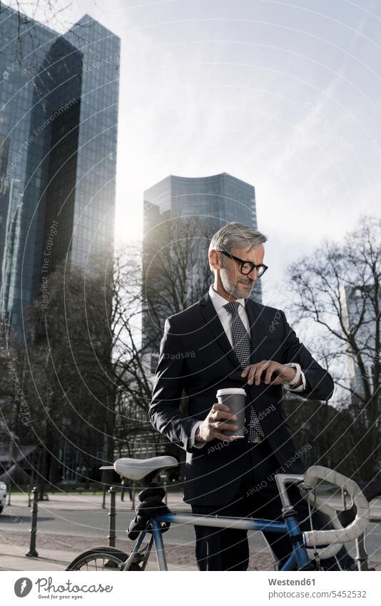 Grauhaariger Geschäftsmann mit Fahrrad und Kaffee in der Stadt, der sich Smartwatch ansieht Bikes Fahrräder Räder Rad staedtisch städtisch Businessmann