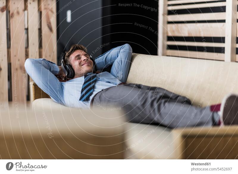 Geschäftsmann mit Kopfhörern auf der Couch liegend in Bürolounge liegt Businessmann Businessmänner Geschäftsmänner Kopfhoerer entspannt entspanntheit relaxt
