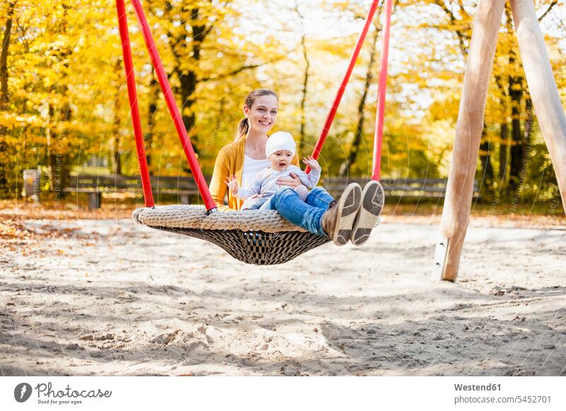 Mädchen, das im Herbst mit seiner Mutter auf einer Schaukel sitzt weibliche Babys weibliches Baby weibliche Babies sitzen sitzend Spielplatz Spielplätze