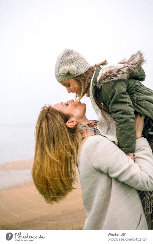 Frau, die im Winter ihre Tochter am Strand hochhebt und küsst Töchter Mutter Mami Mutti Mütter Mama Beach Straende Strände Beaches Kind Kinder Familie Familien