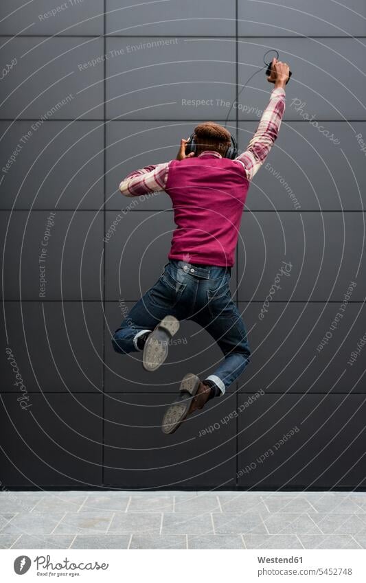Rückenansicht eines glücklichen Mannes, der in die Luft springt, während er mit Kopfhörer und Handy Musik hört springen hüpfen Männer männlich Kopfhoerer Sprung