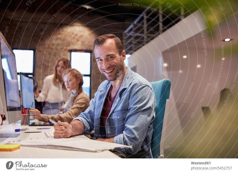 Mann arbeitet am Schreibtisch im Büro und lächelt arbeiten Arbeit sitzen sitzend sitzt lesen Lektüre Office Büros PC Personal Computers Desktop PC schreiben