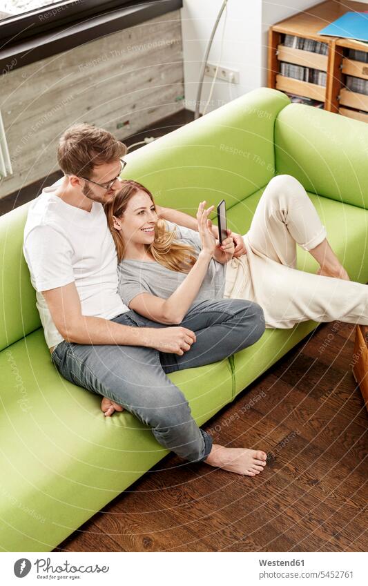 Lächelndes junges Paar auf der Couch im Wohnzimmer zu Hause, das sich ein Handy teilt Pärchen Paare Partnerschaft Mobiltelefon Handies Handys Mobiltelefone
