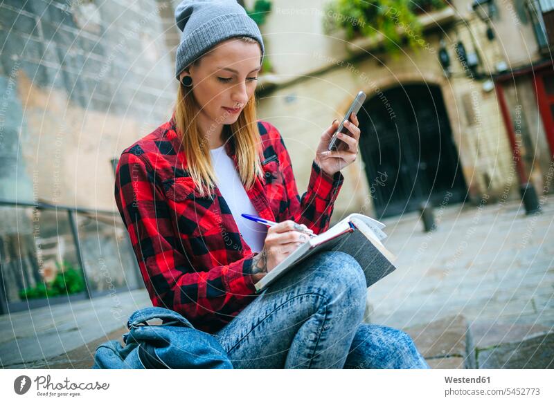 Spanien, Barcelona, junge Frau mit Handy sitzt auf der Treppe und schreibt in ein Notizbuch weiblich Frauen Notizbücher Notizbuecher Erwachsener erwachsen