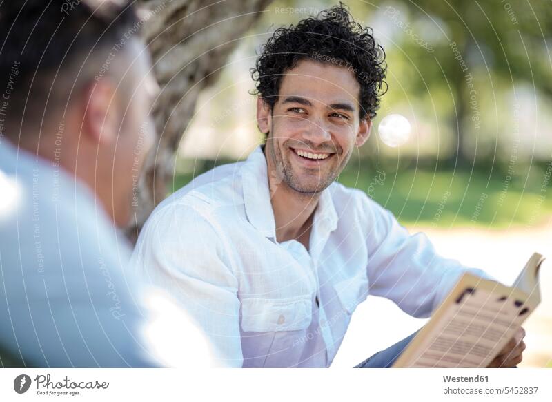 Lächelnder junger Mann mit einem Buch in der Hand schaut einen Freund im Park an lächeln Bücher Männer männlich Erwachsener erwachsen Mensch Menschen Leute