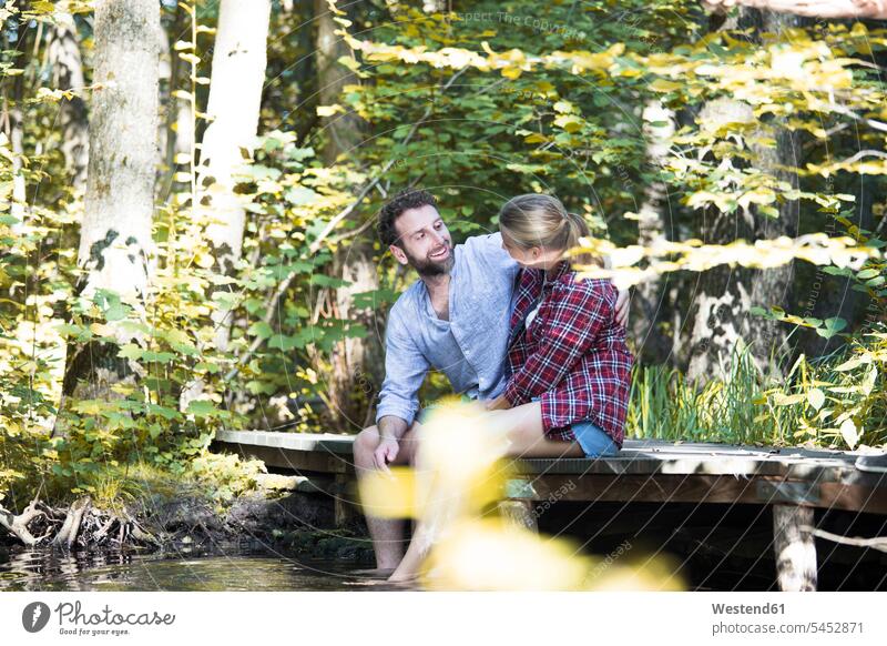 Junges Paar sitzt auf einer Brücke im Wald mit Füßen im Wasser lächeln Pärchen Paare Partnerschaft Bruecken Brücken Bach Bäche Baeche Forst Wälder sitzen