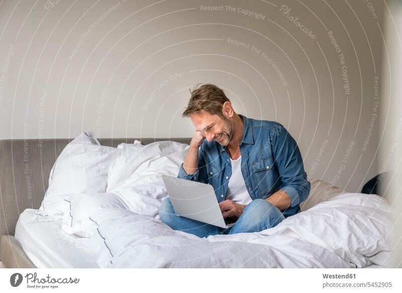 Lächelnder reifer Mann sitzt mit Laptop auf seinem Bett Betten Notebook Laptops Notebooks Männer männlich Computer Rechner Erwachsener erwachsen Mensch Menschen