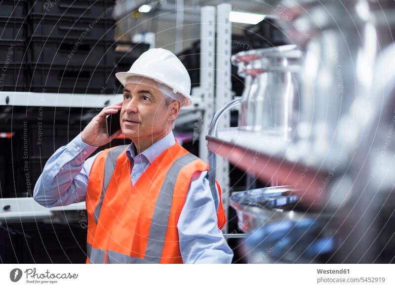 Mann in der Fabrikhalle mit Sicherheitsweste und Schutzhelm, der mit dem Handy telefoniert Männer männlich telefonieren anrufen Anruf telephonieren Mobiltelefon