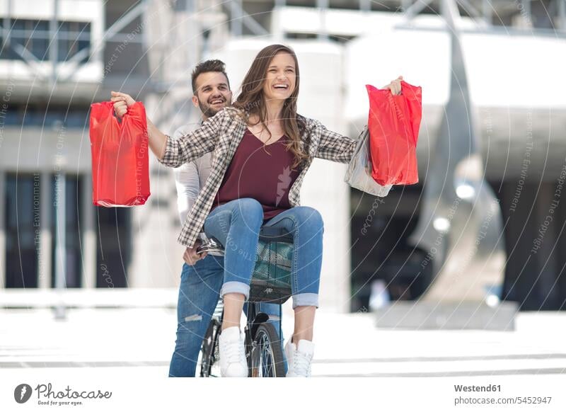 Glückliches junges Paar amüsiert sich in der Stadt beim Fahrradfahren mit Einkaufstaschen Pärchen Paare Partnerschaft lachen shoppen einkaufen Spaß Spass Späße