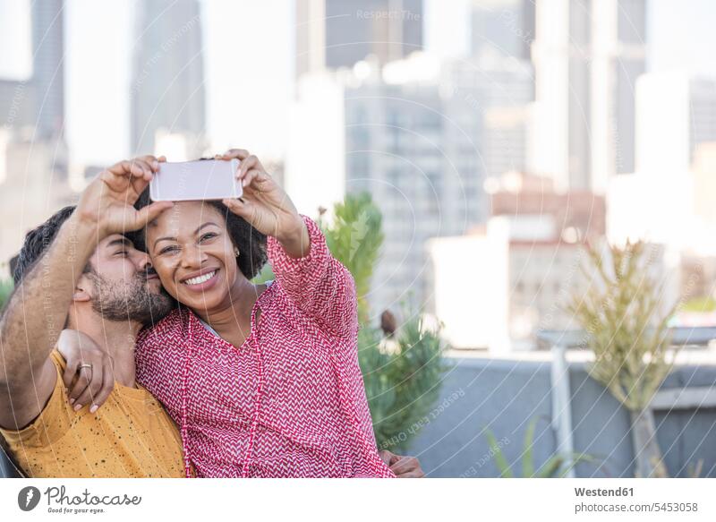 Ein Paar sitzt auf einer Dachterrasse und macht Selfies mit dem Smartphone fotografieren lächeln sitzen sitzend Pärchen Paare Partnerschaft Dachterrassen Mensch
