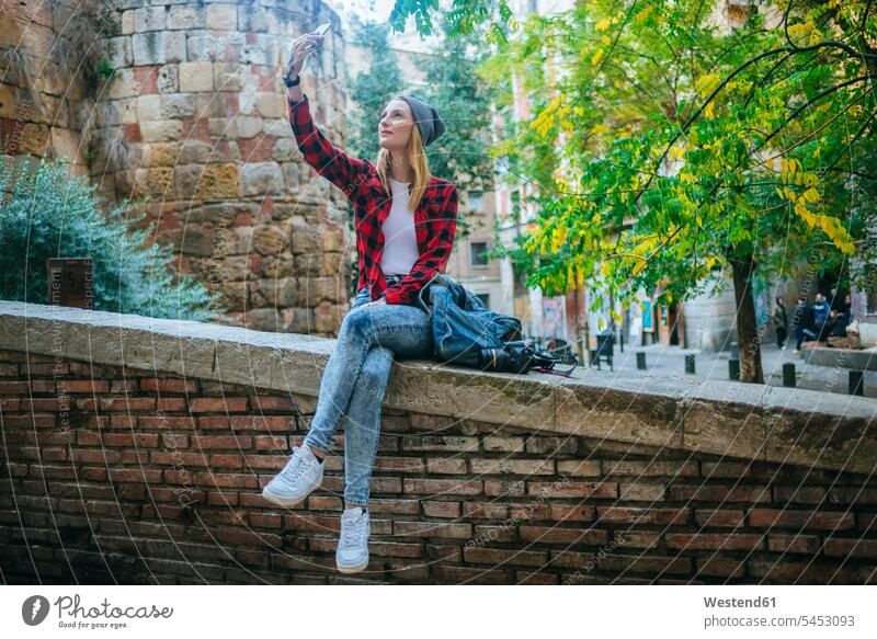 Spanien, Barcelona, junge Frau sitzt auf einer Mauer und macht ein Selfie mit Handy Selfies weiblich Frauen Erwachsener erwachsen Mensch Menschen Leute People